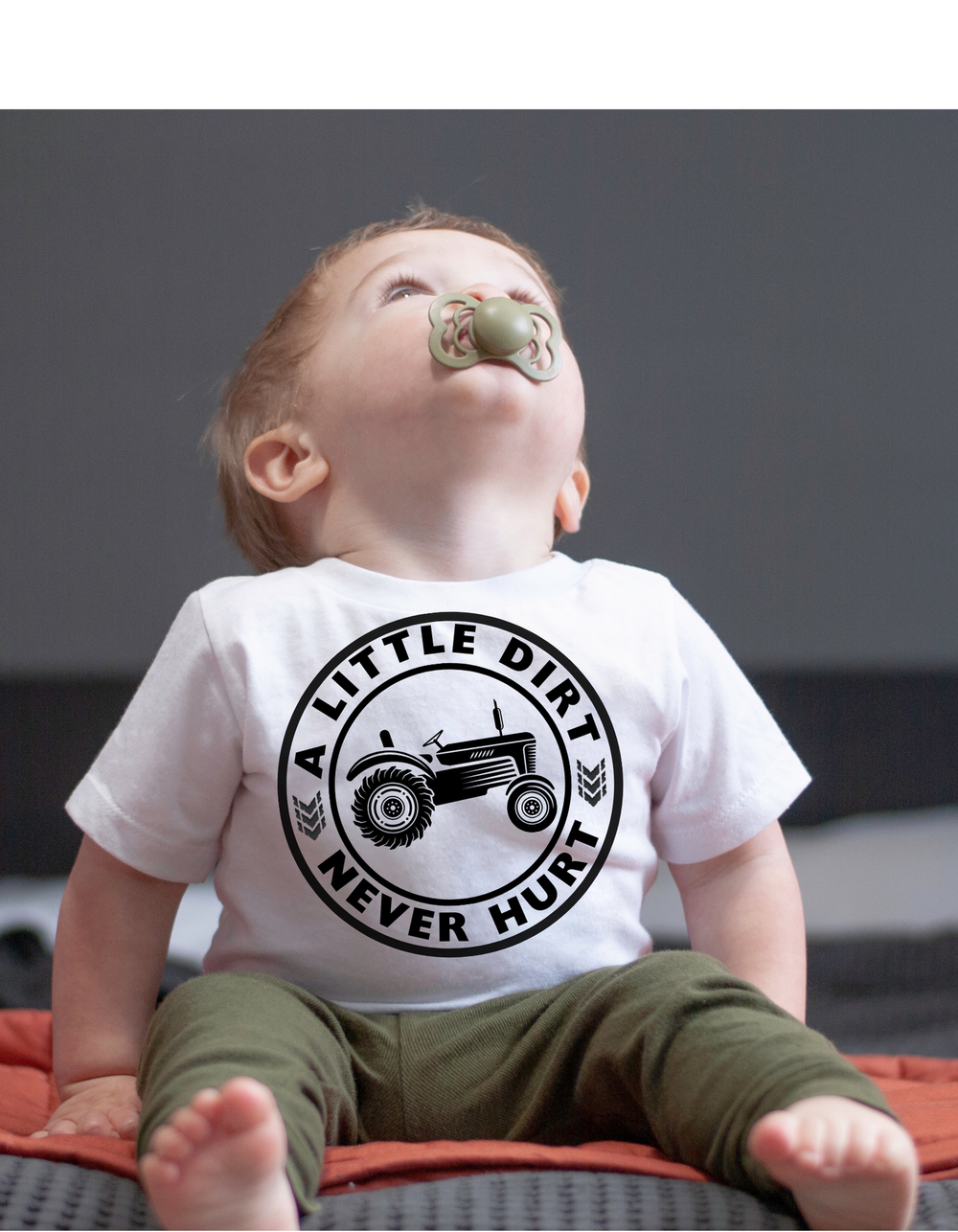 Baby in a white A Little Dirt Never Hurt onesie with a tractor graphic, wearing olive-green pants, sitting on a soft surface with a pacifier.