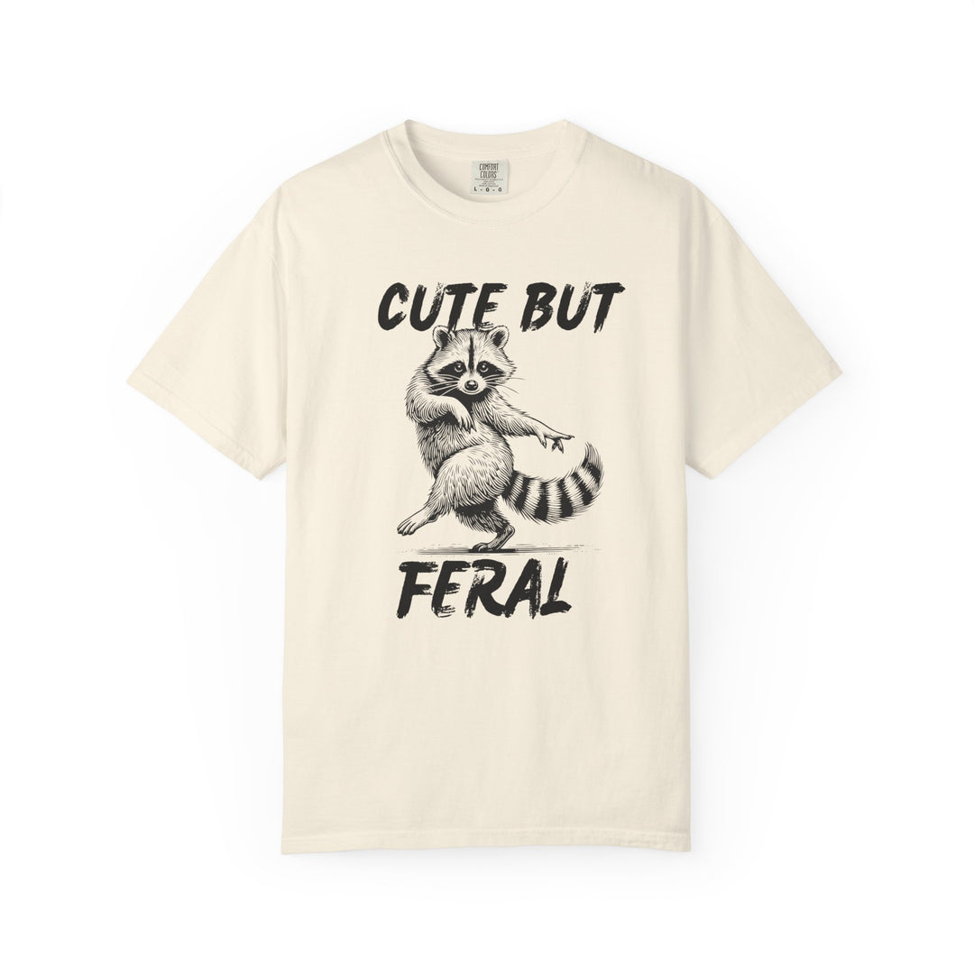 Cute but Feral Tee: Light-colored T-shirt featuring a playful black-and-white raccoon illustration with bold CUTE BUT FERAL text, offering a relaxed, durable fit.