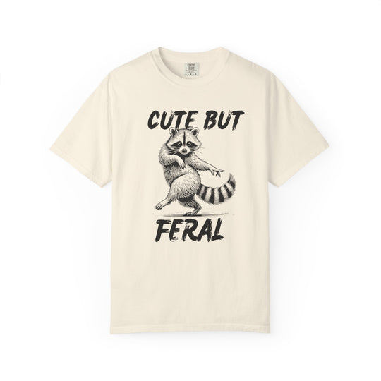 Cute but Feral Tee: Light-colored T-shirt featuring a playful black-and-white raccoon illustration with bold CUTE BUT FERAL text, offering a relaxed, durable fit.