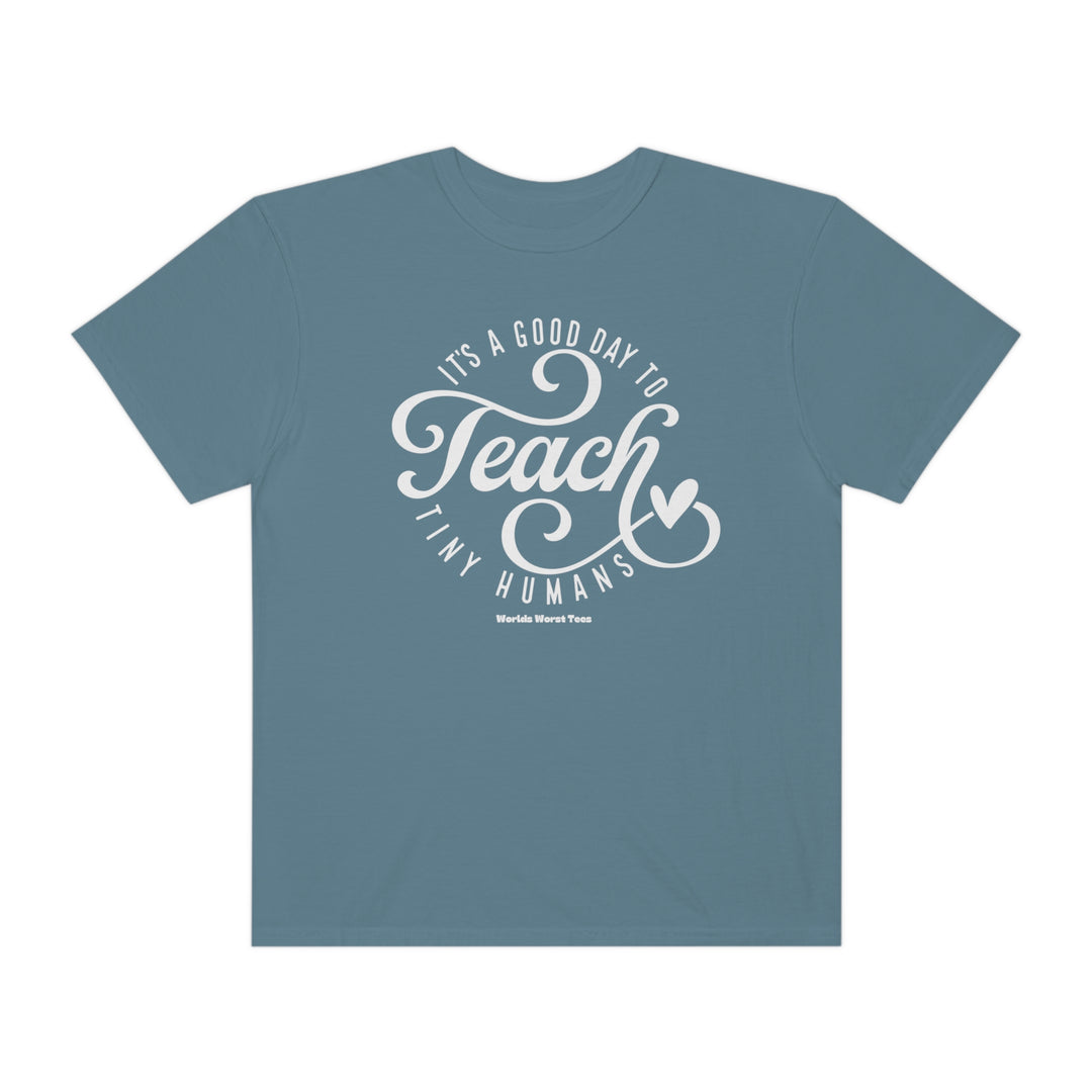 A unisex Teach Tiny Humans Tee in blue with white text. Made of 80% ring-spun cotton and 20% polyester, featuring a relaxed fit and rolled-forward shoulder. Medium-heavy fabric.