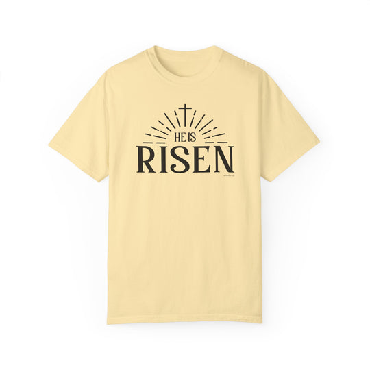 A ring-spun cotton He is Risen Tee in yellow with black text, featuring a cross and rays of light design. Garment-dyed for extra coziness, with a relaxed fit and durable double-needle stitching.
