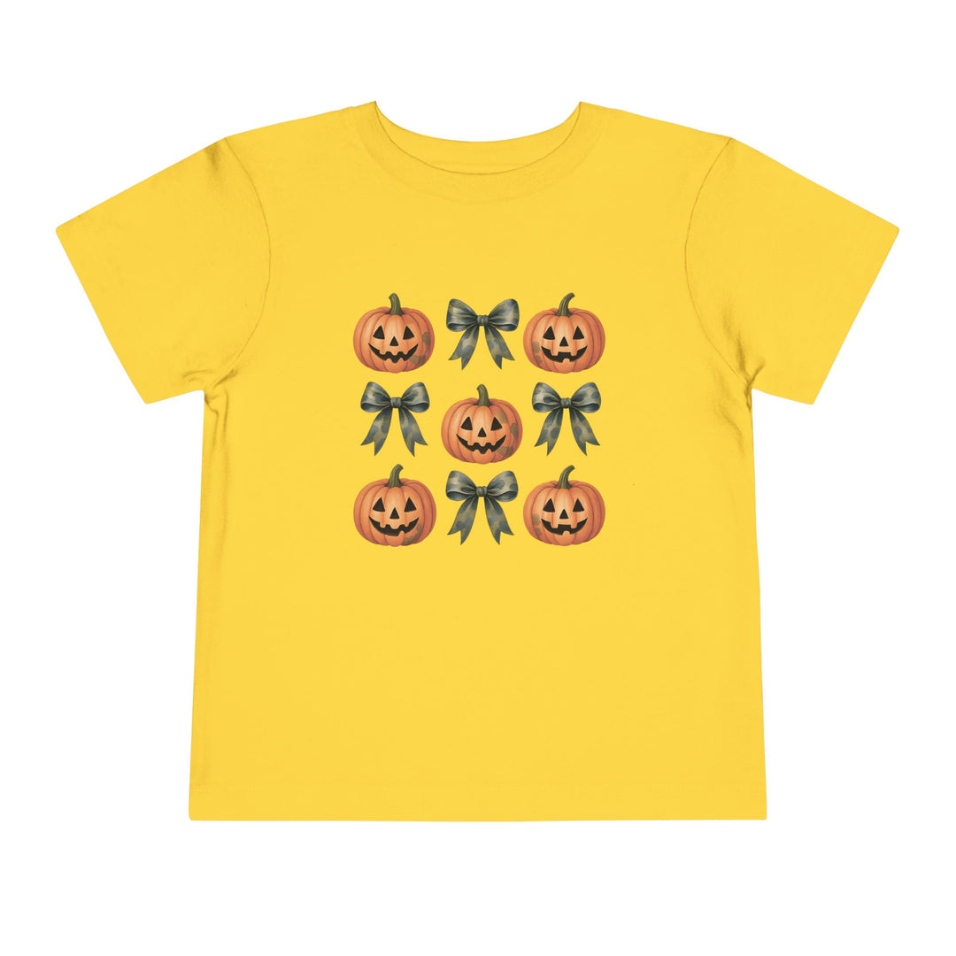 Pumpkins and Bows Toddler Tee features a tic-tac-toe pattern with smiling jack-o’-lanterns and ribbon bows on a bright yellow shirt, perfect for playful, everyday wear.