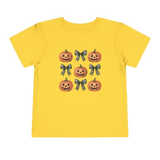 Pumpkins and Bows Toddler Tee features a tic-tac-toe pattern with smiling jack-o’-lanterns and ribbon bows on a bright yellow shirt, perfect for playful, everyday wear.