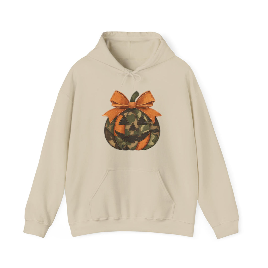 Light-beige Jackie O Hoodie with a woodland-camo jack-o’-lantern graphic, orange bow, kangaroo pocket, cozy cotton-poly blend, unisex fit, perfect for cold days.