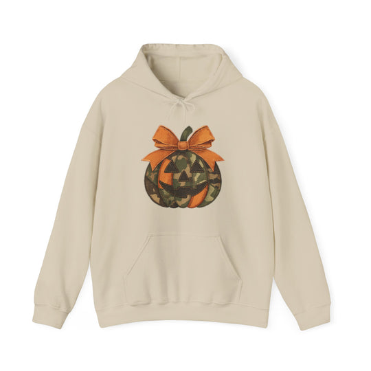 Light-beige hoodie featuring a camouflage and orange jack-o-lantern graphic with a bow, kangaroo pocket, and color-matched drawstring, ideal for cozy, stylish wear.