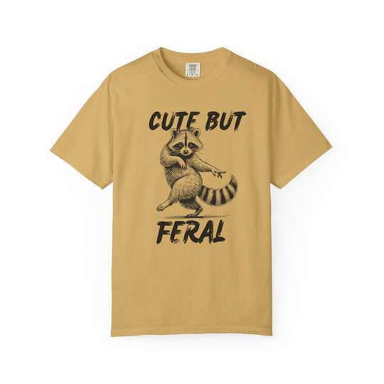 Cute but Feral Tee featuring a playful dancing raccoon illustration and slogan, designed on a mustard-yellow Comfort Colors short-sleeve T-shirt known for its comfort and durability.