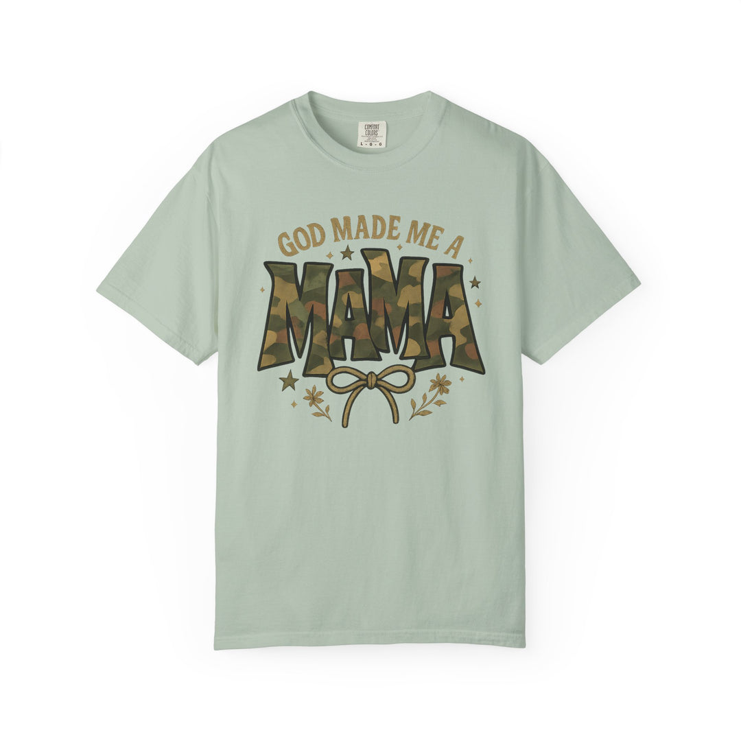 Light sage-green T-shirt with 'GOD MADE ME A MAMA' in camouflage and mustard, featuring a bow and doodles, from Comfort Colors.