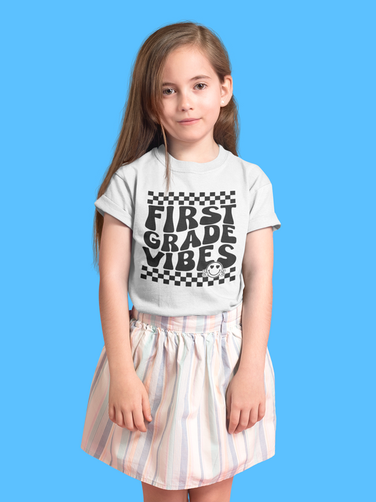 A girl in a white shirt and skirt poses for a picture, showcasing the 1st Grade Vibes Kids Tee from Worlds Worst Tees. This 100% cotton tee features twill tape shoulders and a tear-away label, ideal for everyday wear.