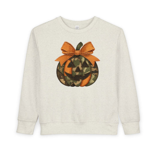 Jackie O Toddler Crew: Cream crewneck sweatshirt with a stylized jack-o’-lantern graphic, featuring orange and camouflage segments, and a large orange bow.