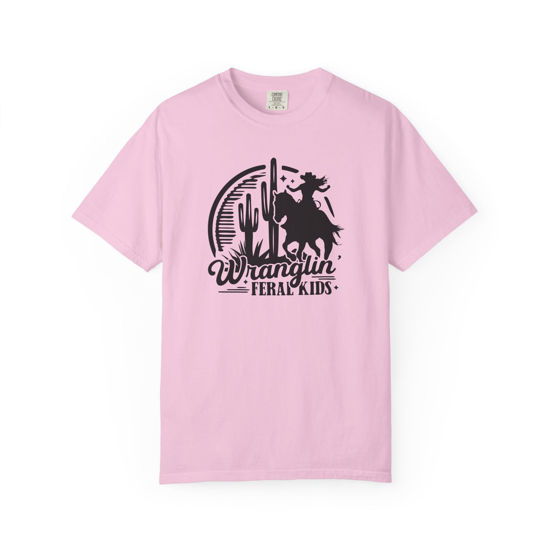Wranglin’ Feral Kids Tee: Light pink Comfort Colors T-shirt featuring a silhouetted rider on a bucking horse, desert scene, and slogan. Available in sizes S to 4XL.
