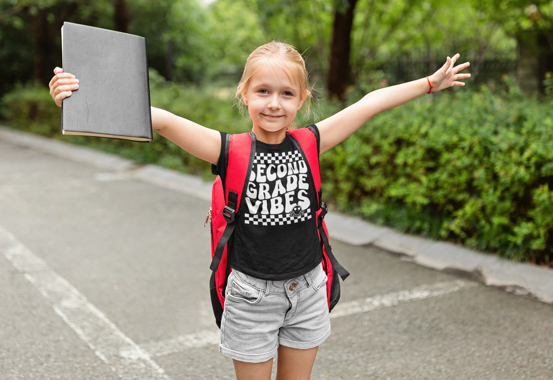 A girl with arms outstretched holds a book, embodying 2nd Grade Vibes Kids Tee. 100% cotton, light fabric, classic fit, tear-away label. Perfect for everyday wear.