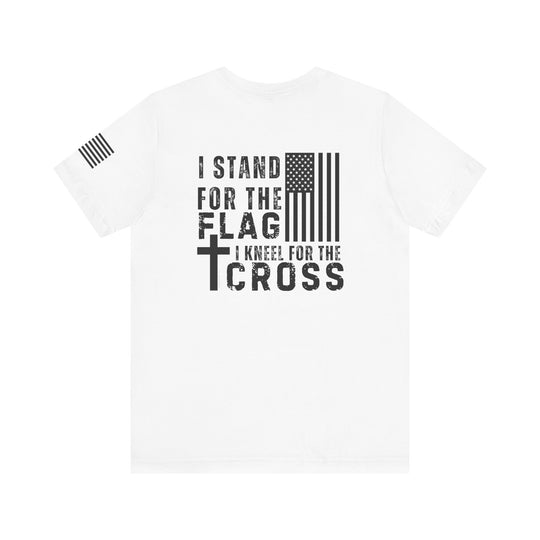 A white Stand for Flag Kneel for Cross Tee with black text and flag design. Unisex jersey tee with ribbed knit collar, 100% Airlume combed cotton, retail fit, tear away label. Sizes XS to 3XL.