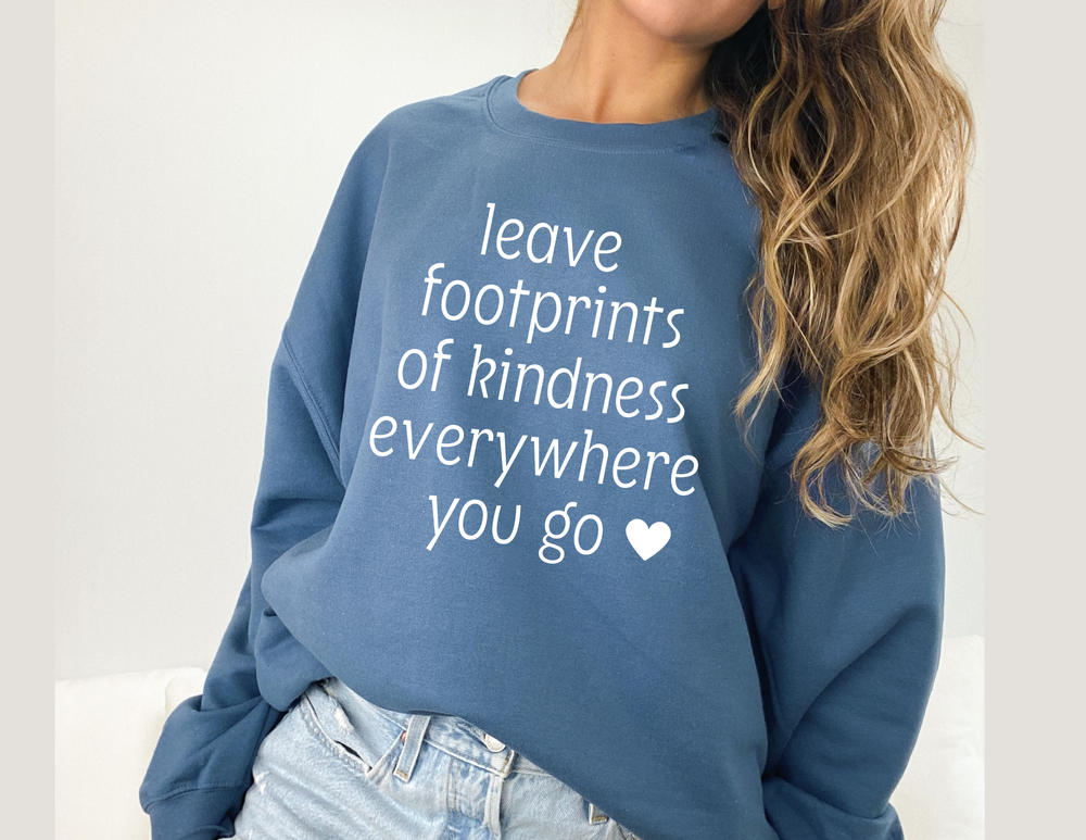 Person wears a slate-blue Footprints of Kindness Crew sweatshirt with white text, paired with light denim jeans, highlighting the cozy, durable design.