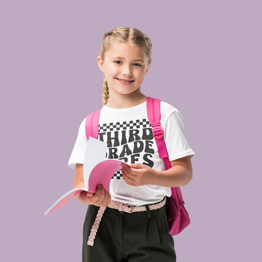 A girl with a backpack holding a book, wearing the 3rd Grade Vibes Kids Tee from Worlds Worst Tees. Features 100% cotton, light fabric, classic fit, tear-away label, and durable twill tape shoulders.