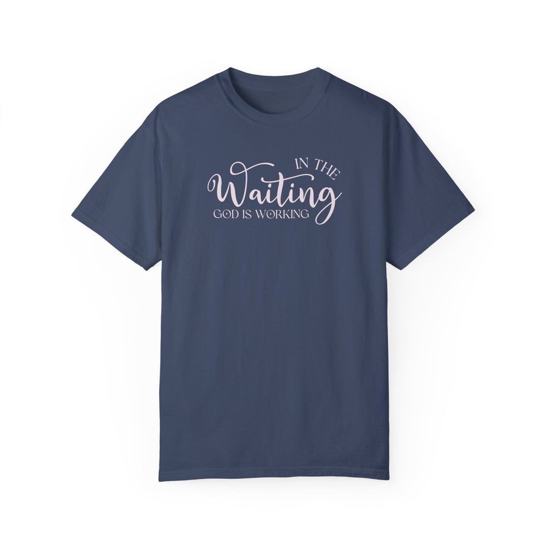 A God is Working Tee, a relaxed-fit, garment-dyed t-shirt in blue with white text. Made of 100% ring-spun cotton for comfort and durability. No side-seams for a tubular shape.