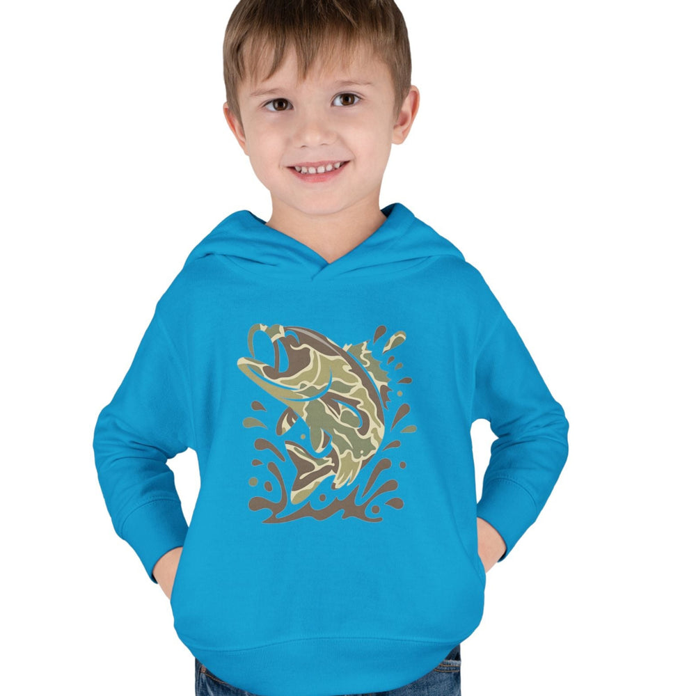 Young boy wearing the Camo Fish Toddler Hoodie, featuring a stylized leaping bass graphic, hands in pockets, smiling against a white background.