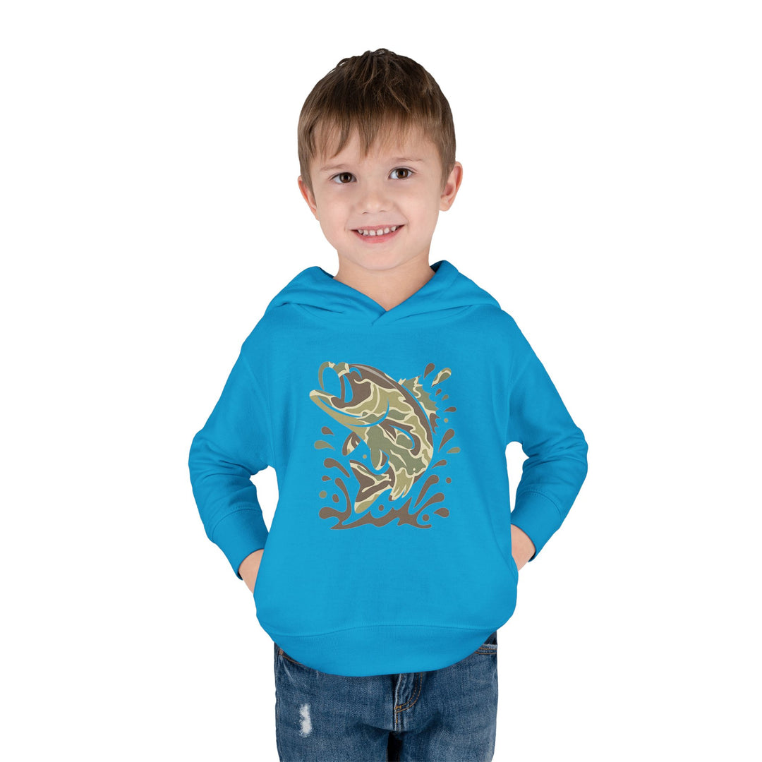 Young boy wearing the Camo Fish Toddler Hoodie, featuring a stylized leaping bass graphic, hands in pockets, smiling against a white background.