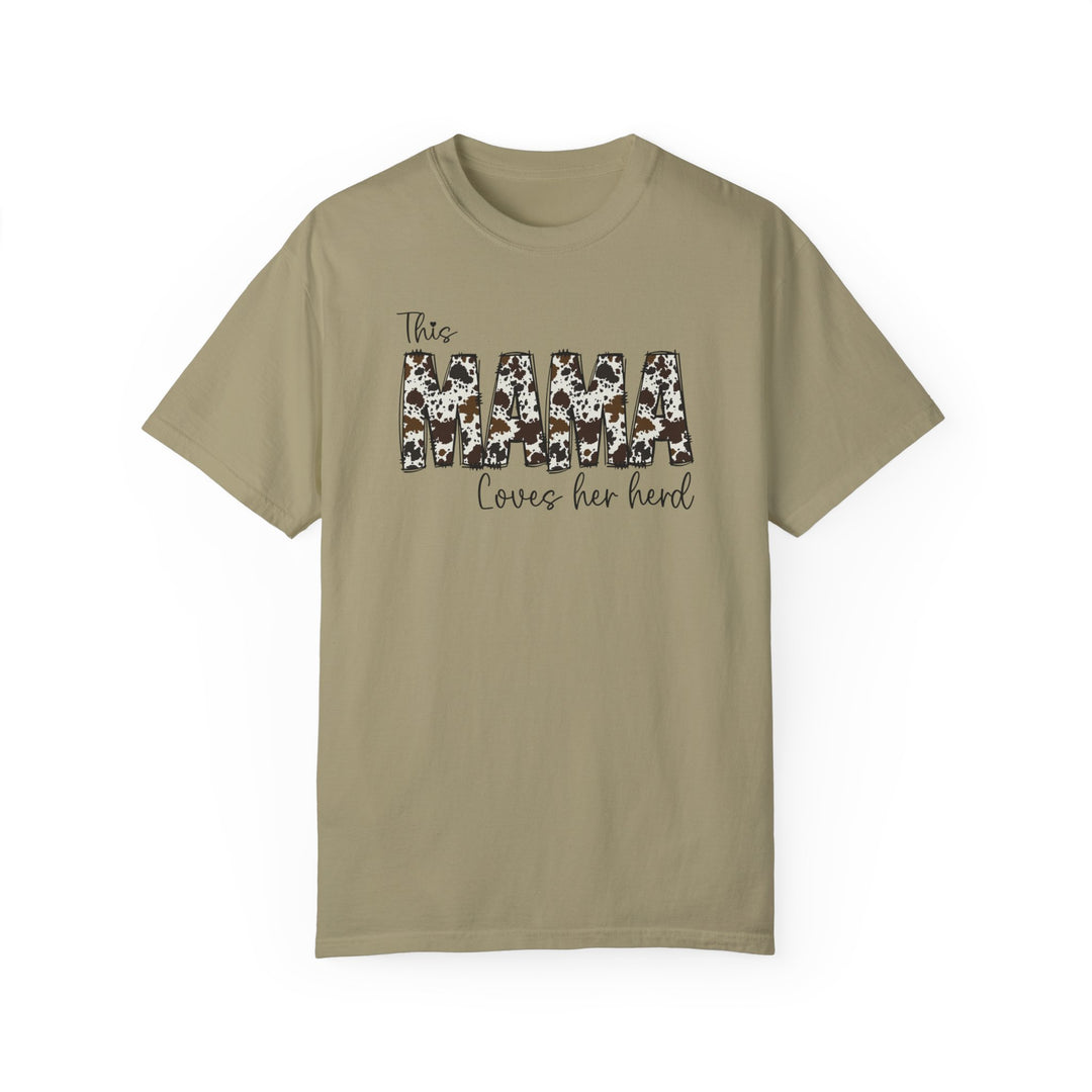 A tan t-shirt featuring a cow print and black text, the Mama Herd Tee. Made of 100% ring-spun cotton, garment-dyed for coziness, with double-needle stitching for durability and a relaxed fit.