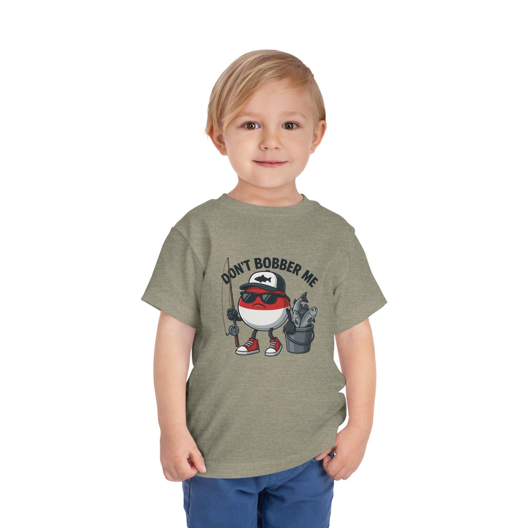 Young blond boy in an olive-green Don't Bobber Me toddler tee with a cartoon fishing bobber character, ideal for playful, everyday wear.
