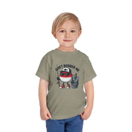 Young blond boy in an olive-green Don't Bobber Me toddler tee with a cartoon fishing bobber character, ideal for playful, everyday wear.