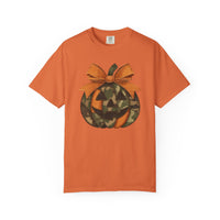 Jackie O Tee features a stylized jack-o’-lantern graphic on a plain orange T-shirt, highlighted by a camouflage-patterned pumpkin and a stitched bow.