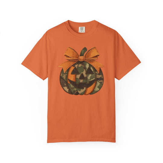 Jackie O Tee features a stylized jack-o’-lantern graphic on a plain orange T-shirt, highlighted by a camouflage-patterned pumpkin and a stitched bow.