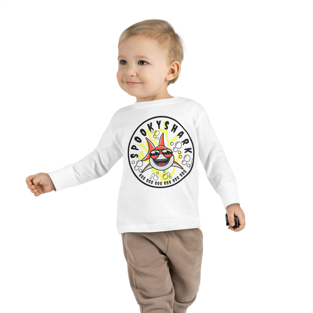A toddler wearing a white long-sleeve tee featuring a cartoon shark with sunglasses. Made of 100% combed ringspun cotton, with ribbed collar and EasyTear™ label for comfort and durability. From 'Worlds Worst Tees'.