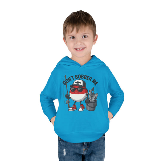 Young boy models the Don't Bobber Me Toddler Hoodie, featuring a cartoon bobber with fishing gear and a playful pun, designed for comfort and durability.