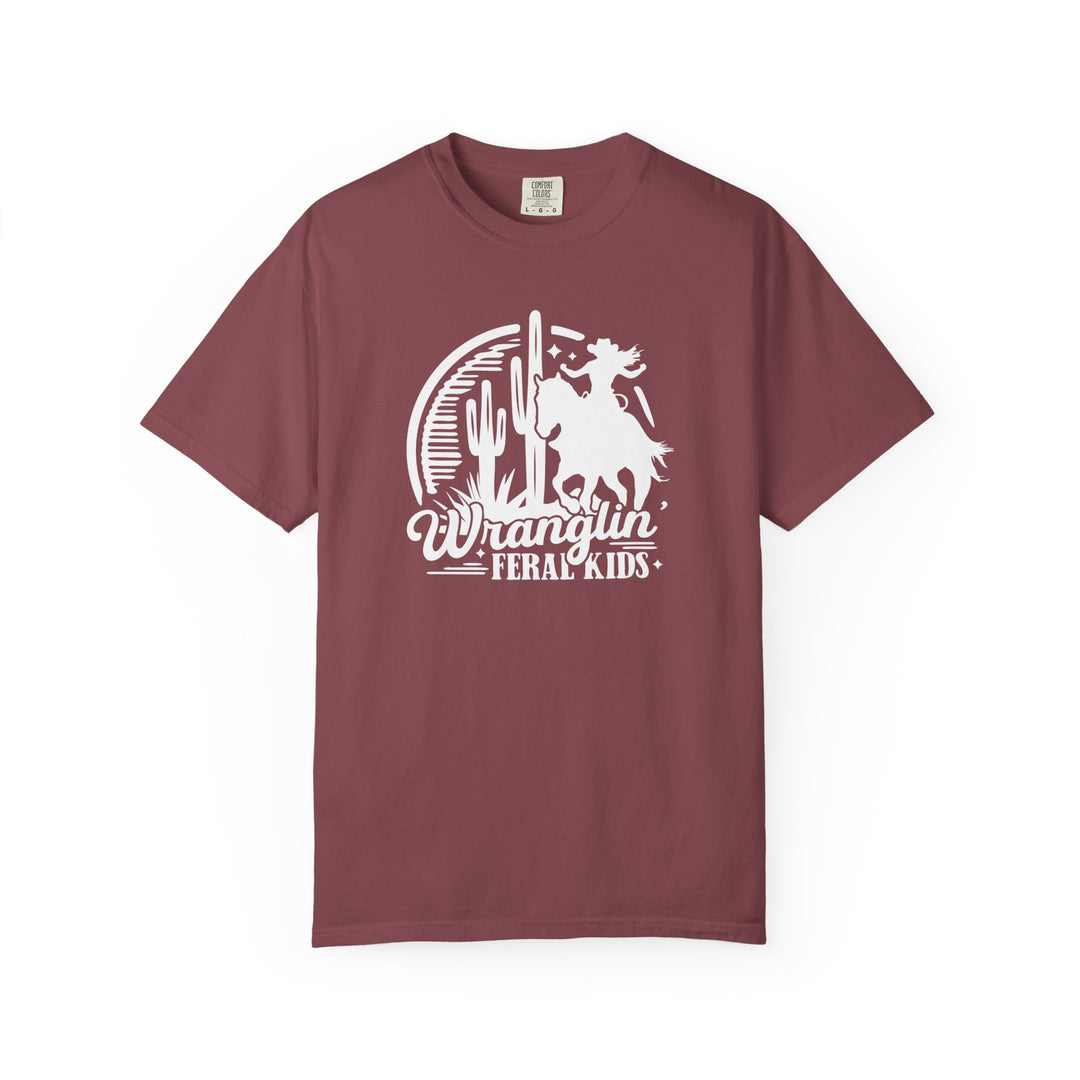 Wranglin’ Feral Kids Tee features a Western desert motif with a cowboy riding past cacti, perfect for a relaxed, durable wear from Comfort Colors.