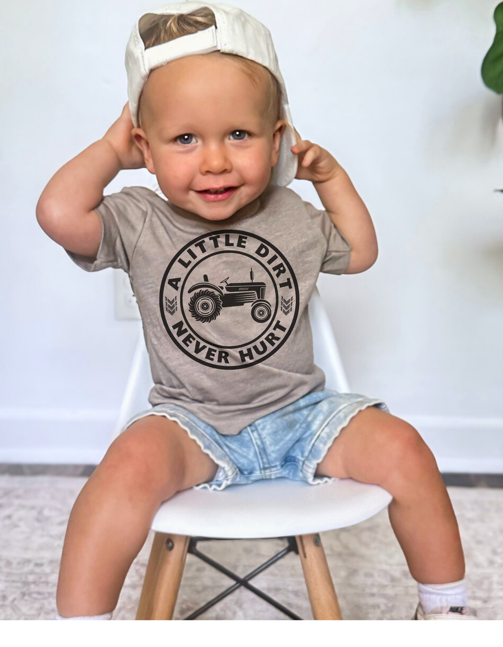 Toddler wearing A Little Dirt Never Hurt tee with tractor graphic, denim shorts, and cap, sitting on white chair. Perfect soft cotton for sensitive skin.