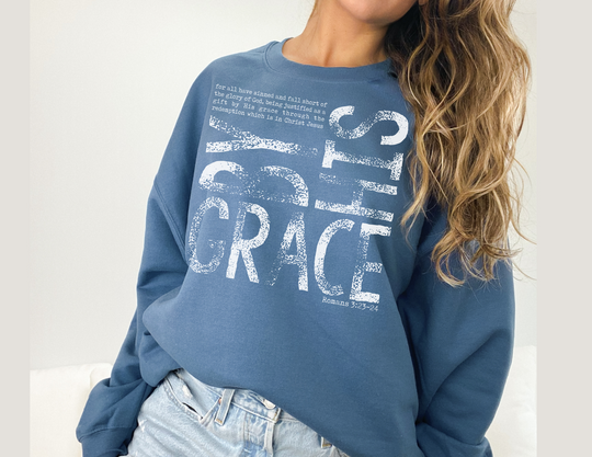 Medium-blue By His Grace sweatshirt with Romans 3:23–24 verse text, paired with light-wash jeans, offering comfort and durability for colder months.