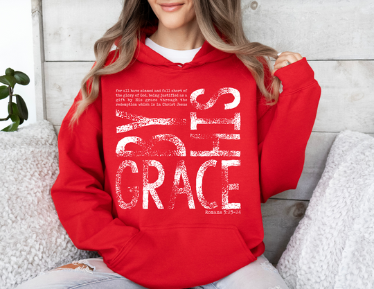 Person in a bright red By His Grace hoodie with Romans 3:23–24 text, seated against a wooden wall, showcasing its cozy, relaxed fit.
