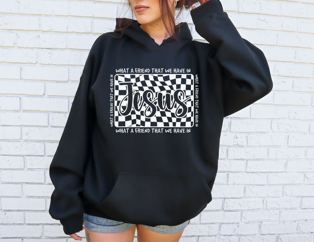 Person wearing an oversized black hoodie featuring a wavy checkerboard graphic with What a Friend We Have in Jesus text, standing against a white-brick wall.