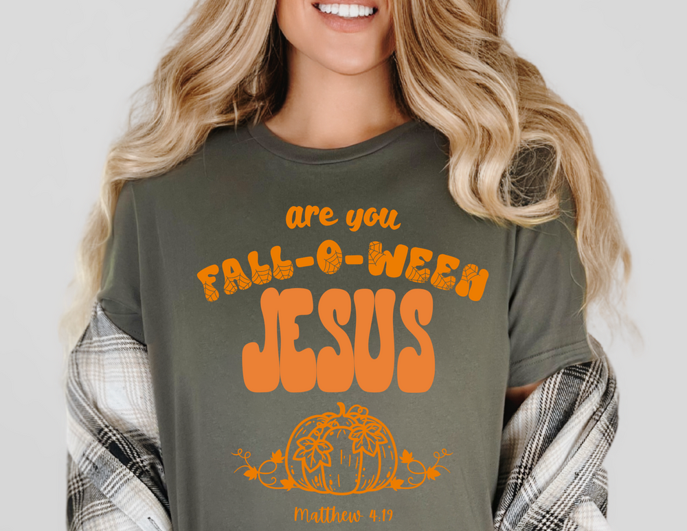 Person wearing a dark-green FALL-O-WEEN Jesus tee with orange lettering, pumpkin graphic, and verse reference. Styled with plaid shirt and wavy blonde hair.