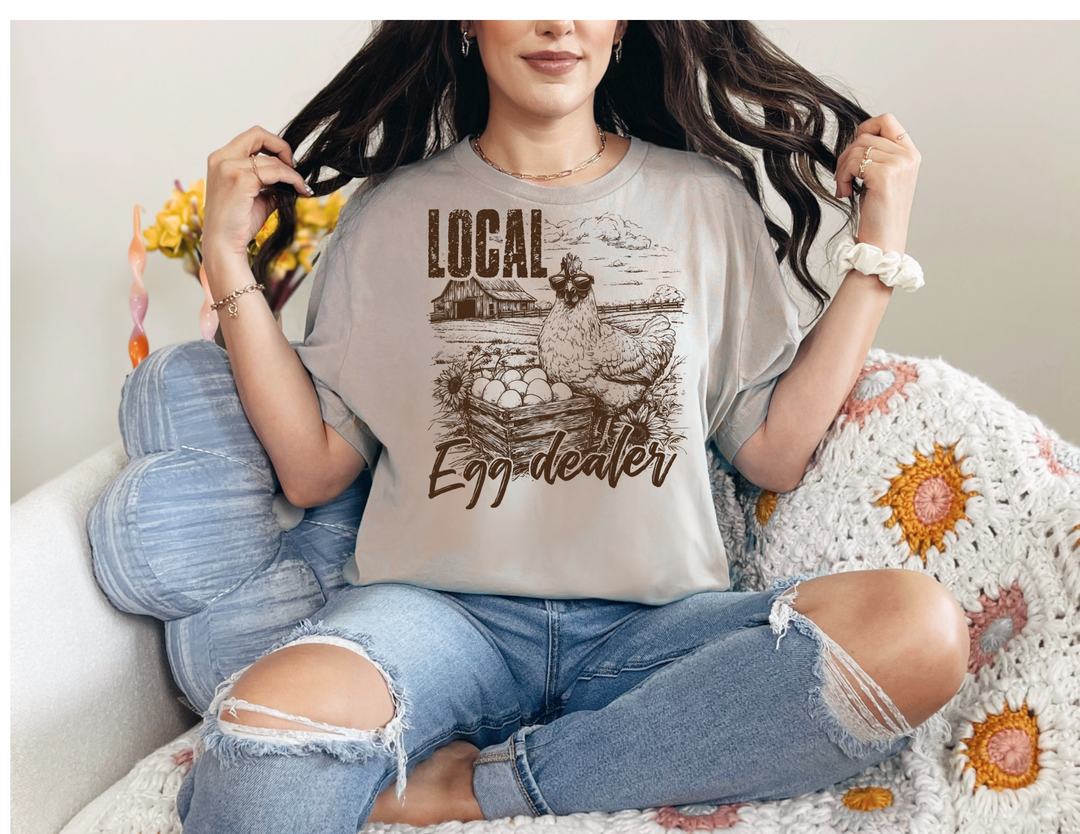The Local Egg Dealer Tee, a classic unisex short sleeve shirt, features ribbed knit collars, tapered shoulders, and durable dual side seams for a lasting fit.