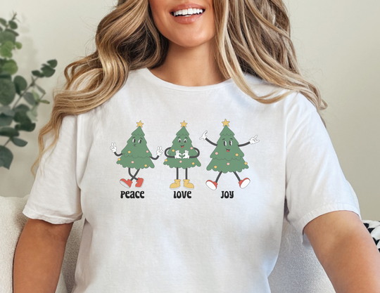 A woman smiling in a Peace Love Joy Tee, featuring a white shirt with cartoon trees. Unisex classic tee made of sustainable US cotton, ideal for casual and semi-formal wear.