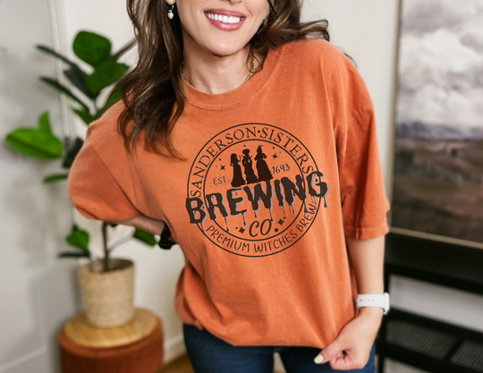 A Sanderson Sisters Brewing Co Tee on a smiling woman in an orange shirt. Unisex Hammer™ T-shirt with classic fit, 100% ring-spun cotton, seamless collar, and double-needle hems. Comfy and stylish.