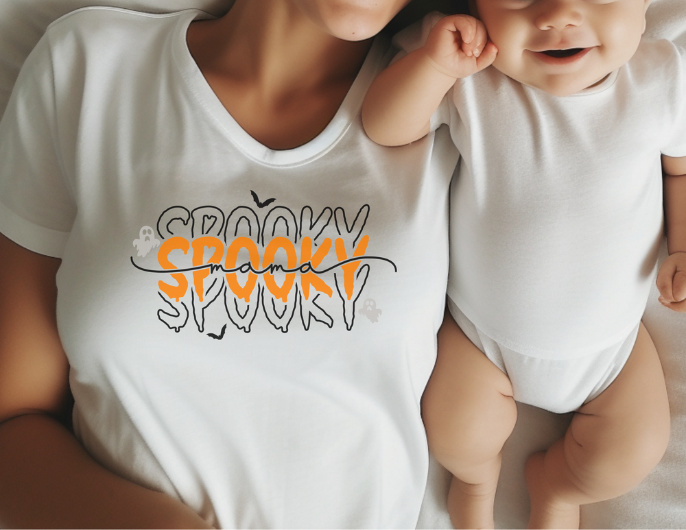 A woman in a white shirt with black text lies on a bed with a baby. The Spooky Mama Tee, made of 100% polyester, offers breathability and style for active days. Perfect for the track or gym.