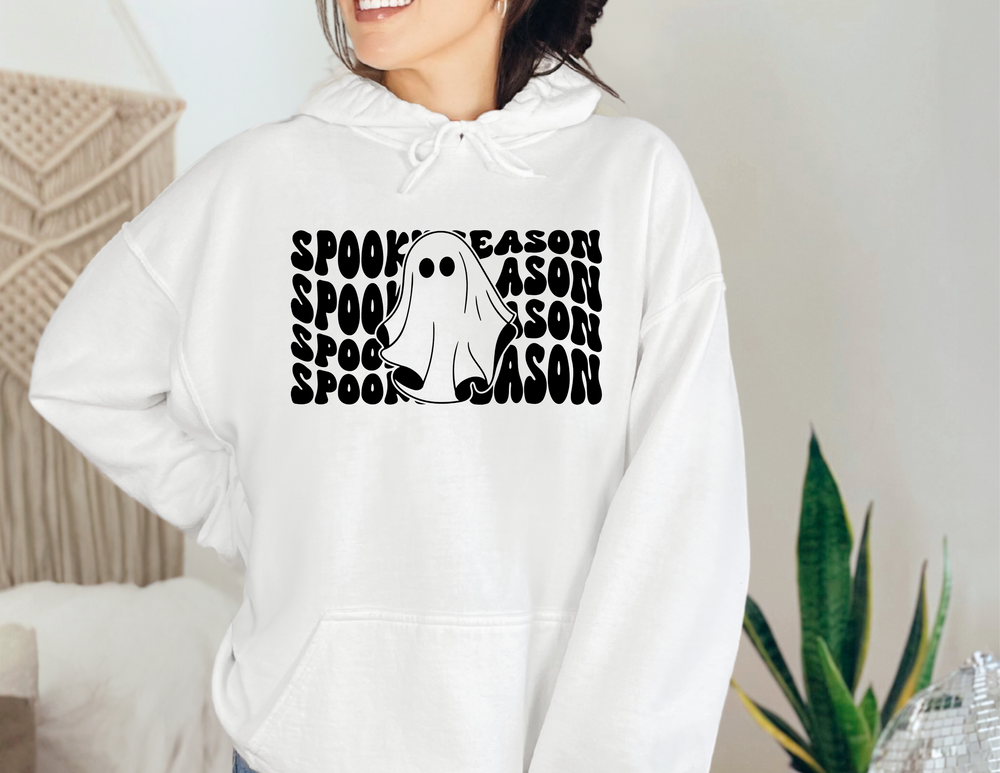 A woman wearing a white Spooky Season Hoodie with a ghost design, made of 50% cotton and 50% polyester. Unisex, plush, and warm, featuring a kangaroo pocket and matching hood drawstring.