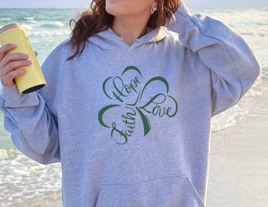 A woman holding a yellow thermos on a beach, showcasing the Faith Hope Love Hoodie from Worlds Worst Tees. Unisex heavy blend, cotton-polyester fabric, kangaroo pocket, and matching drawstring for style and comfort.