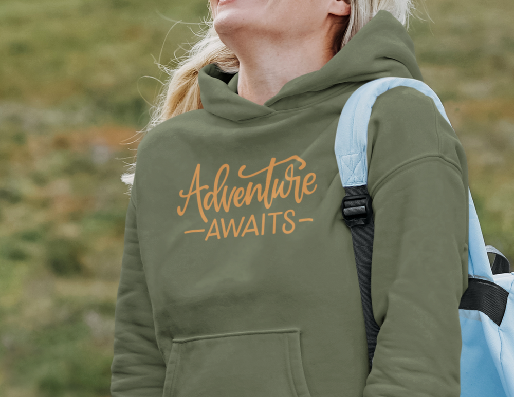 A woman in a green Adventure Awaits Hoodie with a backpack, showcasing a classic fit heavy blend sweatshirt ideal for warmth and comfort. Kangaroo pocket and drawstring hood included.