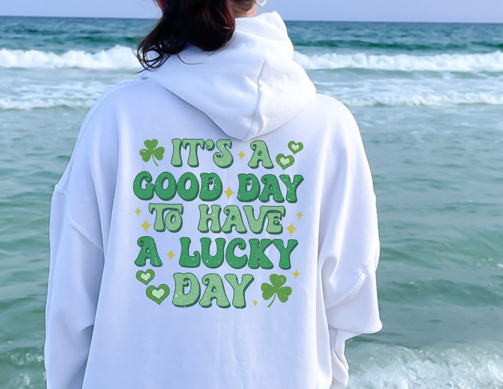 A heavy blend Good Day Lucky Day Hoodie in unisex sizes S-5XL, featuring a person by the ocean, green lettering, and a four-leaf clover design. Cotton-polyester fabric, kangaroo pocket, and matching drawstring.