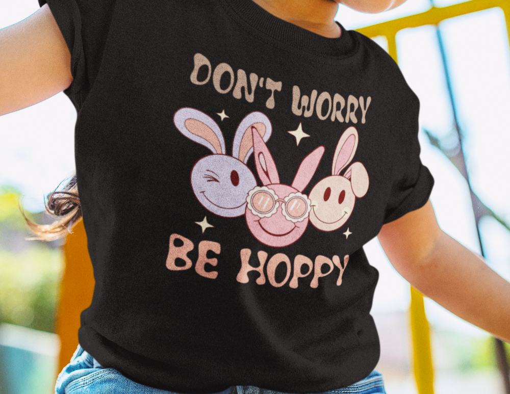 A toddler wearing a black tee with a cartoon bunny, featuring a classic fit for comfort and durability. Made of 100% ringspun cotton, light fabric, and double-needle stitching. Title: Don't Worry Be Hoppy Toddler Tee.