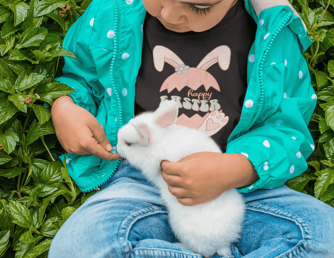 A toddler happily holds a white rabbit on a comfy Happy Easter Toddler Tee. Classic fit, high stitch density, and durable double-needle collar, sleeves, and hems. Made of 100% Ringspun cotton.