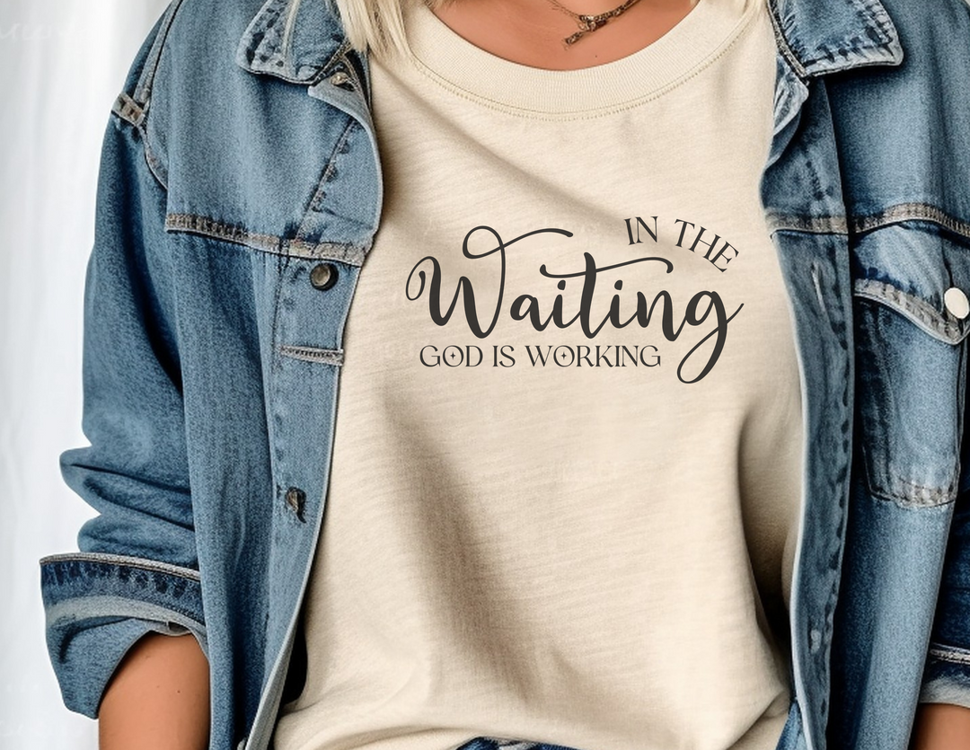 A woman in a denim jacket, showcasing the God is Working Tee from Worlds Worst Tees. A comfortable, ring-spun cotton t-shirt with a relaxed fit and durable stitching, perfect for daily wear.