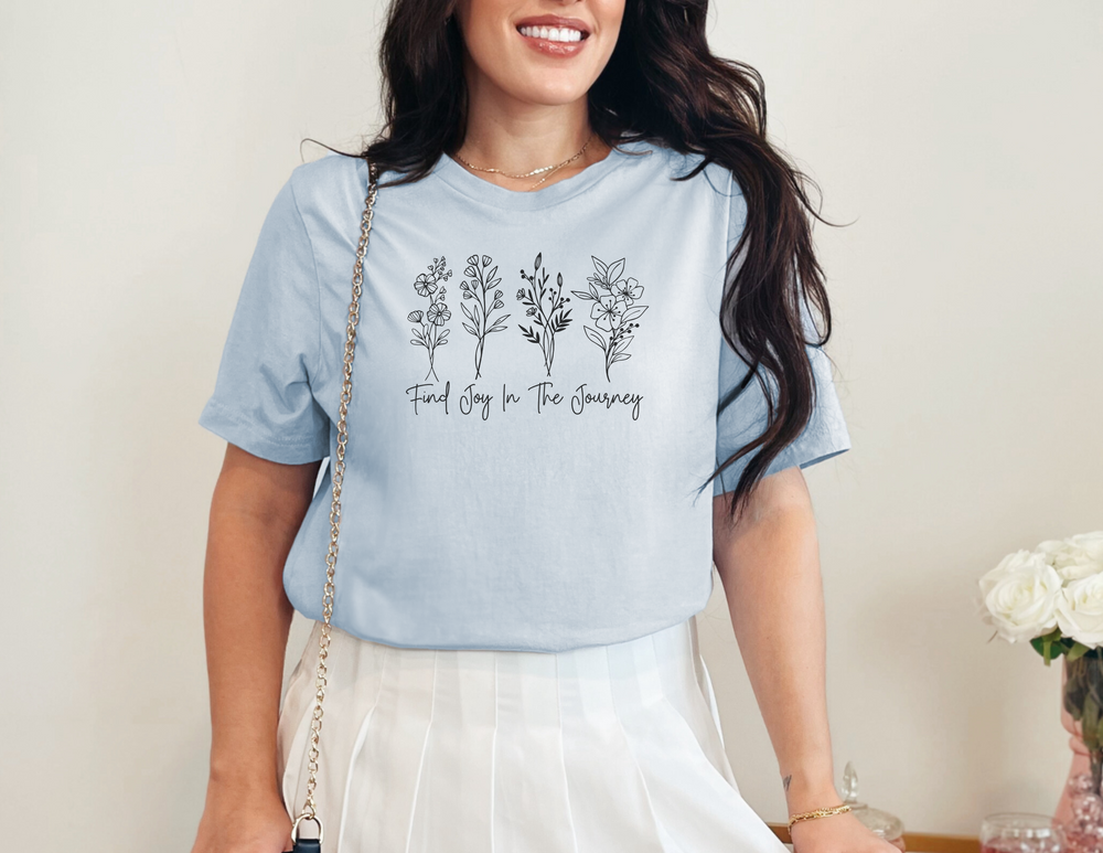 A woman in a blue shirt smiles at the camera, showcasing the Find Joy in the Journey Tee from Worlds Worst Tees. This garment-dyed t-shirt offers a relaxed fit, 100% ring-spun cotton, and durable double-needle stitching.
