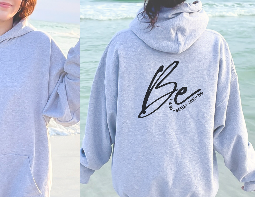A woman in a Be Kind Brave True You Full Zip Hoodie, showcasing a grey sweatshirt with a logo close-up. This medium-heavy, 80% cotton, 20% polyester hoodie offers warmth and style.