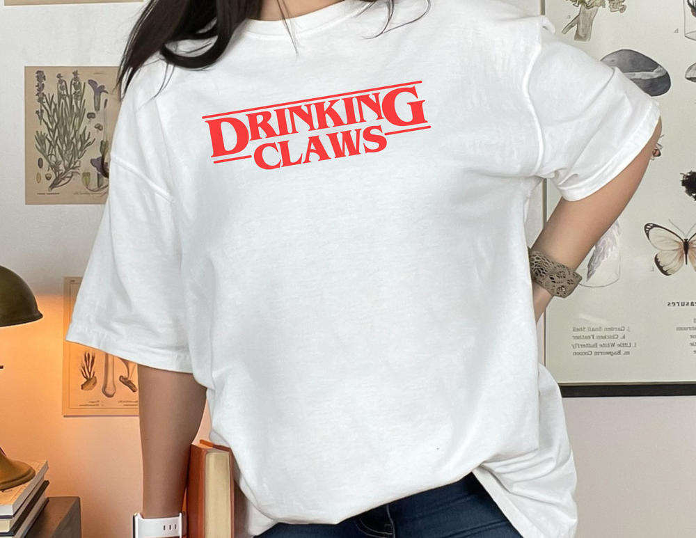 A woman in a white DRINKING CLAWS shirt, showcasing a relaxed fit, ring-spun cotton fabric. Garment-dyed for extra coziness, with double-needle stitching for durability. From Worlds Worst Tees.