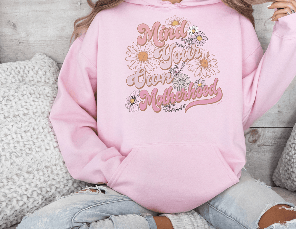 A person in a pink Mind Your Motherhood Hoodie, sitting on a couch. Unisex heavy blend sweatshirt, cotton-polyester mix, cozy and warm. Kangaroo pocket and matching drawstring. Reflects Worlds Worst Tees store ethos.