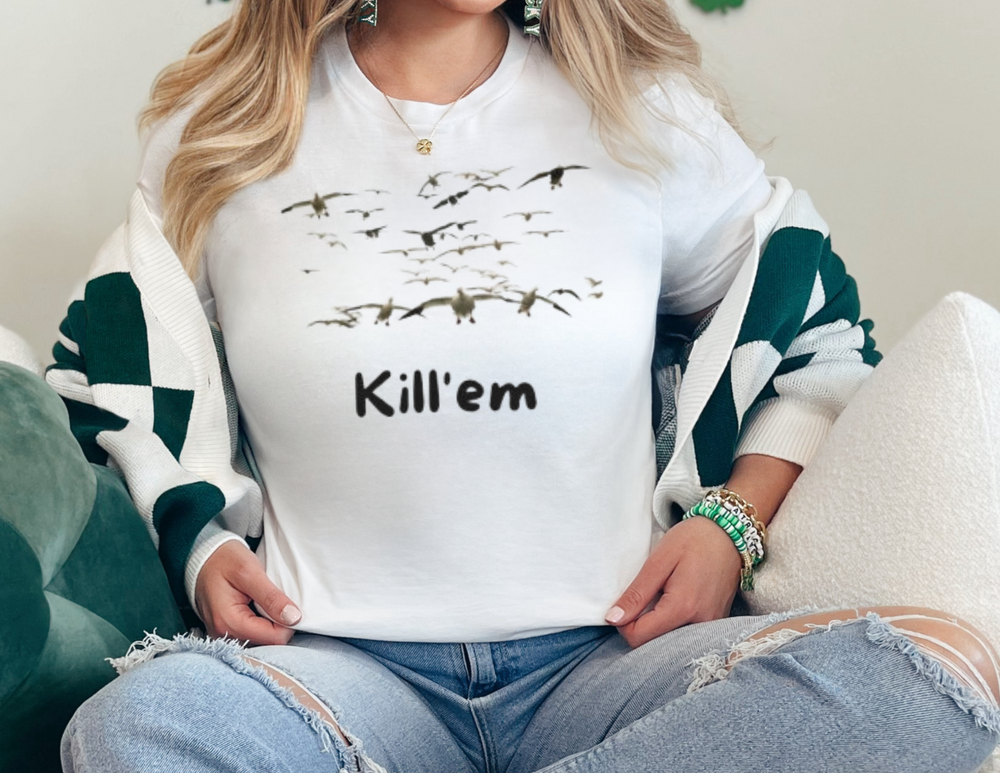 A woman in casual dress sits on a couch, showcasing the Kill'em Snows Tee. Unisex heavy cotton tee with no side seams, ribbed knit collar, and medium weight fabric. Classic fit, true to size.